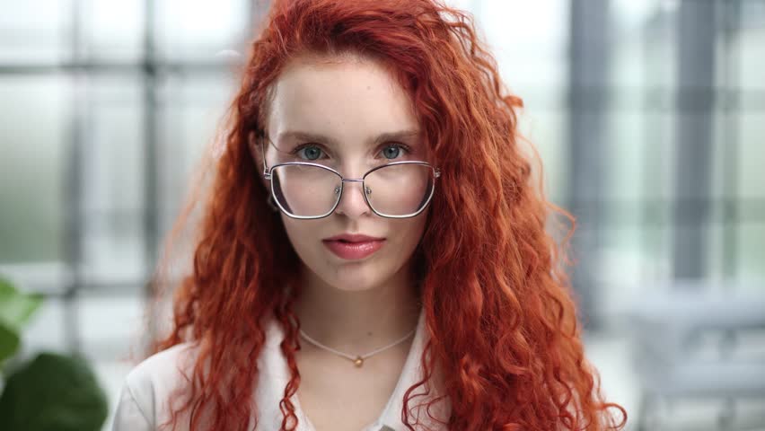 A woman with red hair is wearing glasses and holding them up to her face