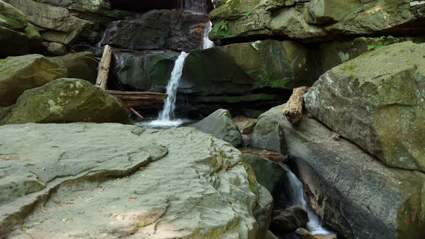A drone view ascending on a multi-tiered waterfall flows through a rocky grotto. Sunlight streams in from above, illuminating the water and stones.