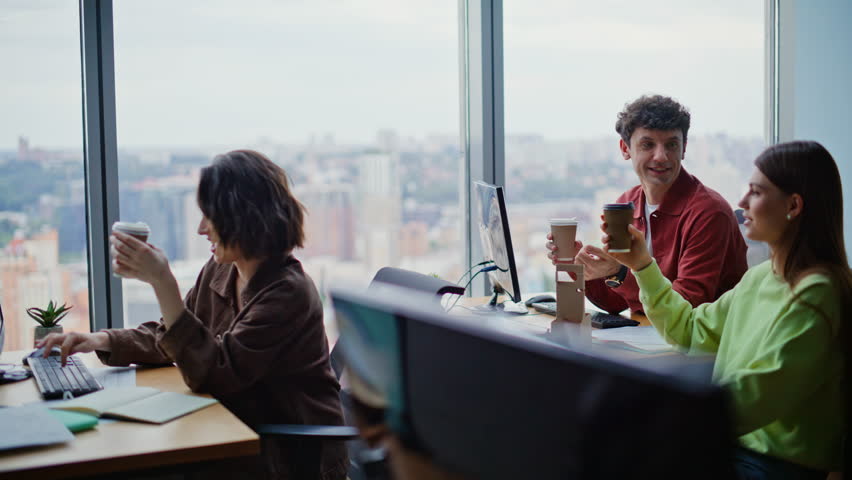 Positive colleagues toasting coffee at work break. Cheerful office coworkers having fun joking together in open space room. Happy young business team enjoying friendly communication enjoying beverages