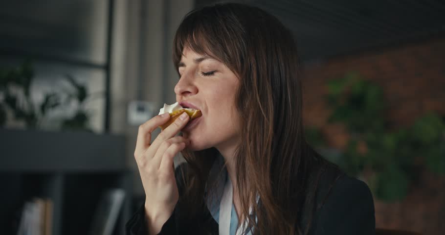 Woman in business attire savoring a delicious pastry with eyes closed enjoying the taste in a modern office setting with natural light and plants around