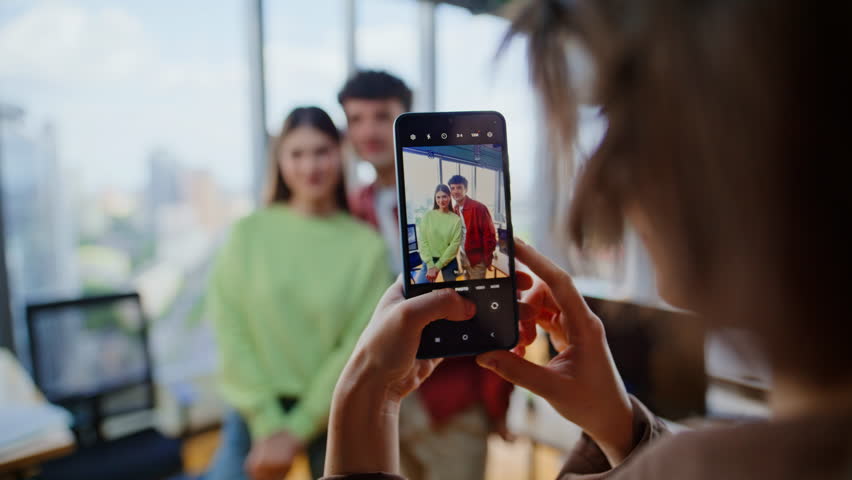 Mobile phone display showing cheerful coworkers posing together near big windows office closeup. Happy young colleagues capturing group selfie photo smiling brightly. Excited people checking snapshot
