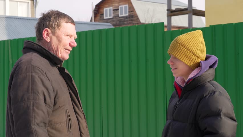Candid outdoor interaction between older man and youth by vivid green barrier on bright winter day