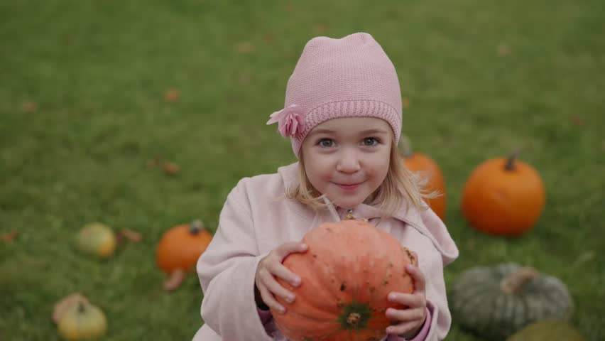 Young girl enjoys the fall season while posing with a pumpkin in the grass surrounded by squash.
