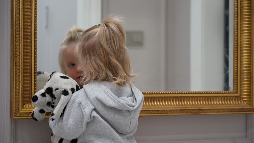 Blonde tot with spotted plush pal joyfully explores her curious reflection in ornate antique gold mirror, pressing her face against glass