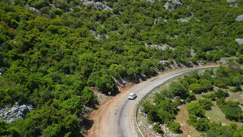 Drone captures white car on bendy Turkish road. Mountains and forests enclose the way. Scenic Turkey, or Turkiye, offers peaceful drives through wild green landscapes.