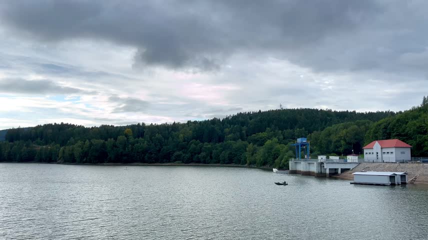 Static video of a boat sailing on a calm lake with forest and buildings in background.