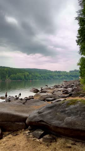Close view of rocky lakeshore under dark storm clouds. Gentle rain creates ripples on calm water surface.