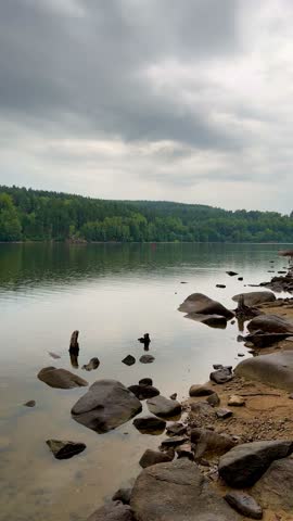 Static video of a rocky shore and calm lake surface with raindrops falling, forest in background under cloudy sky.