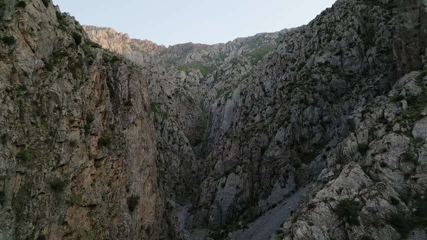 Morning drone flyover reveals a steep Uzbekistan gorge, limestone wall and a dark ravine with sparse alpine greenery. Soft dawn light touches the ridge and invites adventure in the Chimgan region