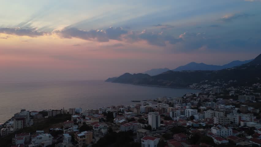 Aerial drone shot of a coastal town in Montenegro during sunset. The sun casts a golden reflection over the Adriatic Sea as the drone moves right and backwards, revealing rooftops and scenic coastline