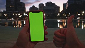 Person Holding Smartphone with Green Screen and Giving Thumbs Up Gesture Against Blurry City Night Lights, Perfect for Technology and Social Media Concepts - Powered by Shutterstock - Get 15% off with code: PIKWIZARD15