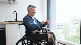 Senior Caucasian man sitting in wheelchair by kitchen indoors using phone near window. Scene depicts everyday lifestyle, accessibility, comfort, connection, communication, independence, and inclusion - Powered by Shutterstock - Get 15% off with code: PIKWIZARD15