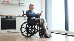 Senior Caucasian man sitting in wheelchair by kitchen indoors using phone near window. Scene depicts everyday lifestyle, accessibility, comfort, connection, communication, independence, and inclusion - Powered by Shutterstock - Get 15% off with code: PIKWIZARD15