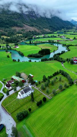 Breathtaking vista of Stryn, Norway showcasing vibrant green fields and meandering rivers. The serene landscape is framed by majestic mountains and shrouded in mist, creating a tranquil atmosphere.