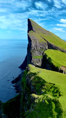 Breathtaking cliffs rise dramatically over the ocean in the Faroe Islands as visitors explore the lush green terrain. a couple at Mylingur cliff hiking trail