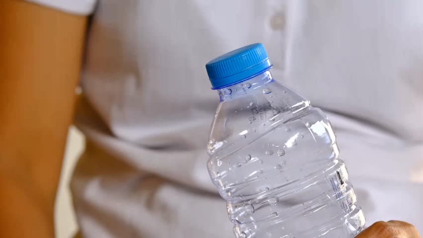 Closeup view 4k stock video footage of 500 ml transparent plastic bottle full of fresh water closed with blue plastic cap isolated on grey wall background. Woman opens bottle