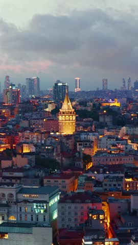 Vertical view of the historic Galata Tower standing out among the illuminated rooftops and modern skyscrapers of Istanbul's skyline at sunset