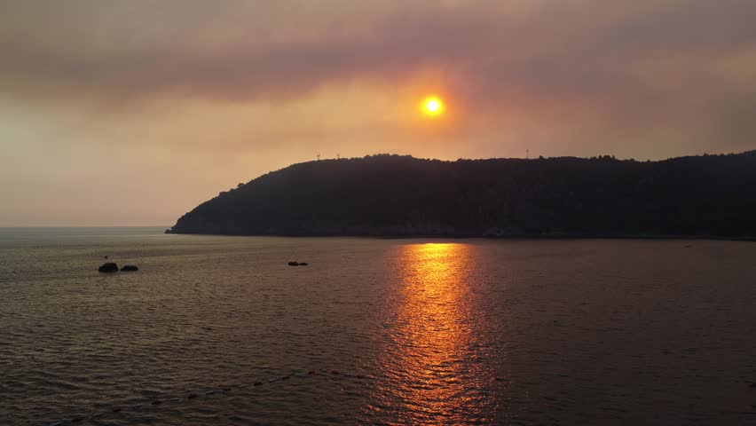 Aerial drone video of sunset over Adriatic Sea in Montenegro, with orange sunlight reflection on water and soft haze in the sky. Scenic coastal view with dramatic evening light as drone moves upward.