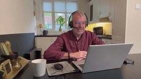 Senior man with headset microphone on video call using laptop computer at kitchen table. Remote work meeting and digital communication for mature adults at home. - Powered by Shutterstock - Get 15% off with code: PIKWIZARD15