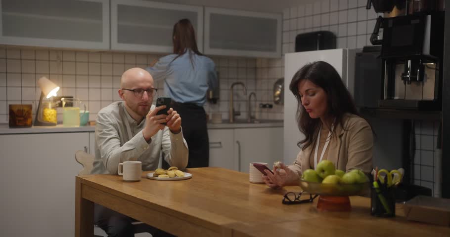 Office workers guy and girl sitting at the table having lunch and looking at social networks on the phone instead of chatting in the office kitchen. Busy during break