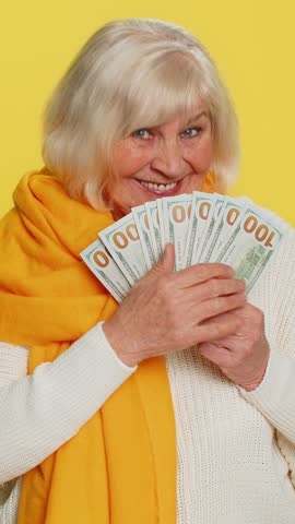 Rich happy senior old woman waving money dollar cash banknotes bills like a fan, success business career, lottery winner, big income wealth pension. Elderly grandmother pensioner on yellow background