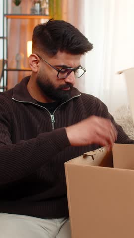Indian man at home opens delivery box, sees incorrect item and shakes head in disappointment. Hispanic guy on sofa looks upset and frustrated by online shopping mistake and order fulfillment issue