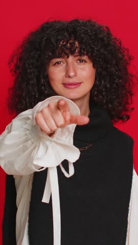 Hey you. Happy Caucasian young woman with curly hair smiling excitedly and pointing forward to camera, choosing lucky game winner, indicating to awesome you, promotion. Girl isolated on red background