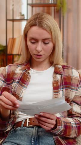 Young adult woman holds bills on home sofa shakes head in disappointment after reviewing high expenses. Blonde girl reacts with frustration, feeling concerned about financial pressure and rising costs