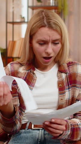 Young adult woman holds bills on home sofa shakes head in disappointment after reviewing high expenses. Blonde girl reacts with frustration, feeling concerned about financial pressure and rising costs
