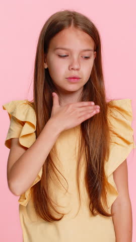 Young child girl with tired sad expression coughs into elbow, showing signs of sickness physical discomfort. School kid isolated on pink background, feeling weak worried about health and well-being