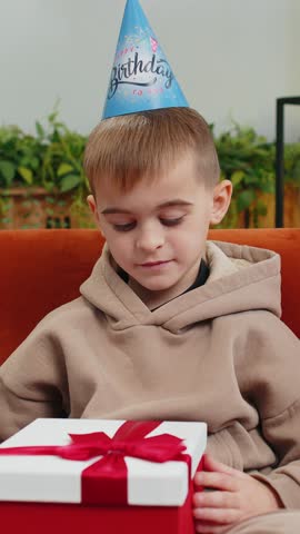 Happy preteen child boy wears festive birthday cap hat hold gift box with ribbon congratulating. Teenager male little kid celebrating party event opening delivery greeting present at home room on sofa