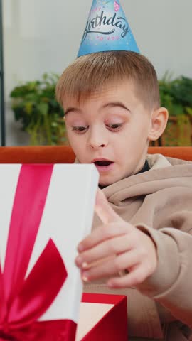 Happy preteen child boy wears festive birthday cap hat hold gift box with ribbon congratulating. Teenager male little kid celebrating party event opening delivery greeting present at home room on sofa