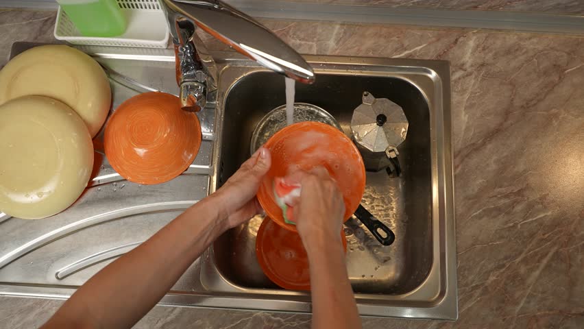 Close-up side view of a woman washing plates, scrubbing and rinsing a bowl with soap suds under the tap. A woman is washing dishes with a sponge and foam