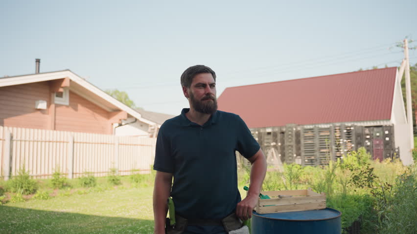 Young bearded man in dark polo stands posed in sunny backyard beside blue drum with wooden crate and garden tools, farmhouse and fence behind, calm confident look