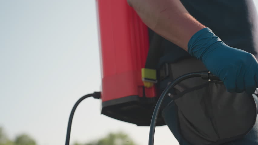 close up of man fumigating field with backpack sprayer wearing glove and protective gear, sunlight highlighting arm and hose during outdoor agriculture pest control