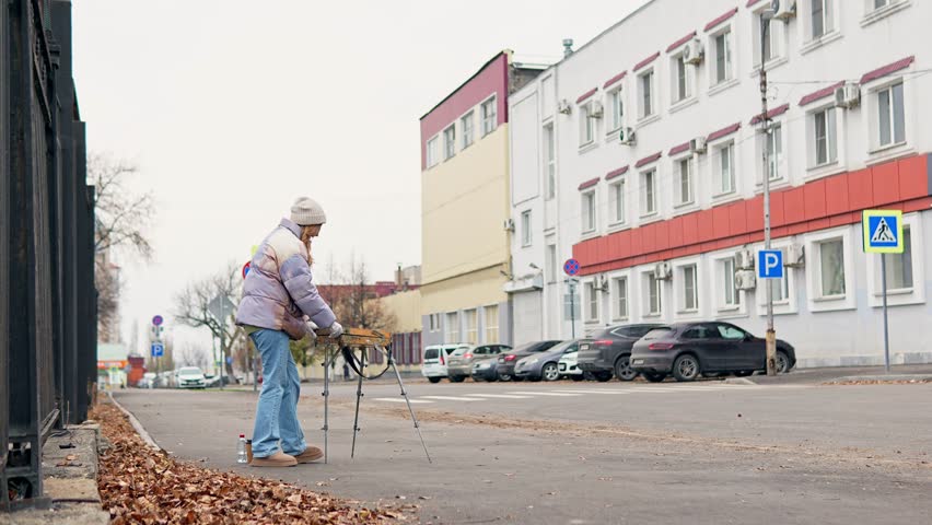 Scenery of woman in jacket and hat opening portable art kit and arranging supplies filmed in slow motion. She is measuring proportions with finger vision method preparing to paint a picture outdoors