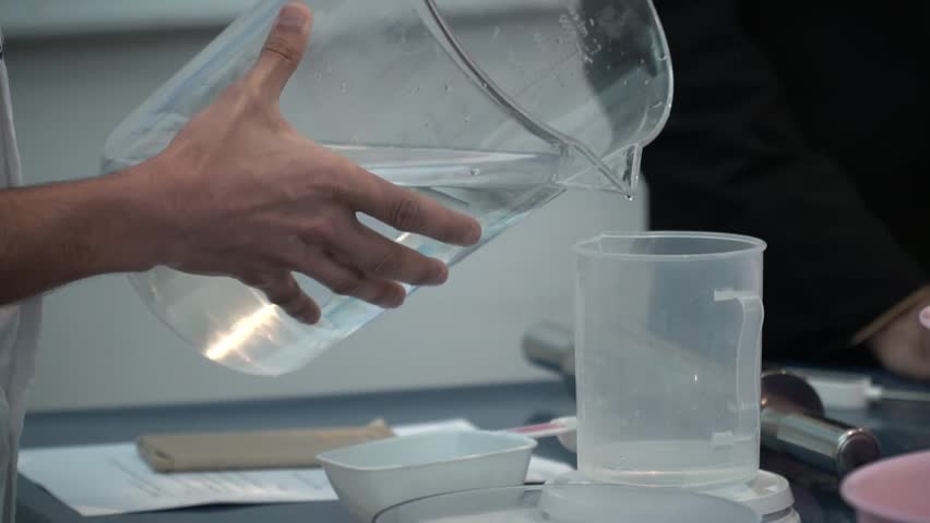 A person pouring water from a measuring jug into a container on a table.