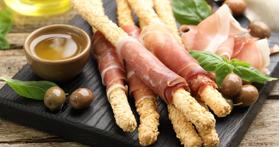 Woman taking grissini breadstick with prosciutto from board at wooden table, closeup