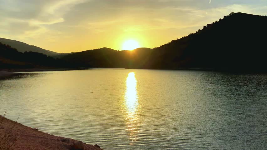 Romantic sunset scene from Lago di Fiastra in the Sibillini Mountains, minutely rippling waters mirroring the light of the sun, shining behind the silhouettes of the surrounding peaks, dramatic sky