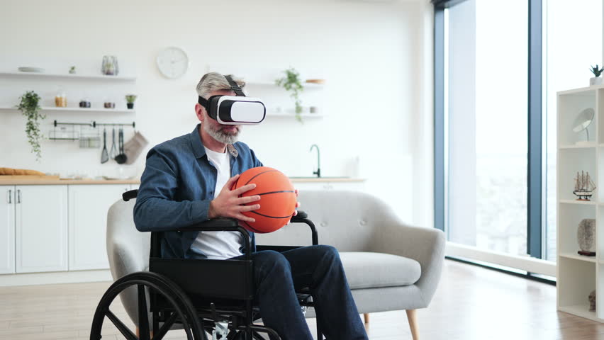 Senior man sitting in wheelchair indoors wears VR headset holding basketball, enjoying technology. Interior setting includes sofa, shelves. Emphasizes virtual reality, accessibility - Powered by Shutterstock - Get 15% off with code: PIKWIZARD15