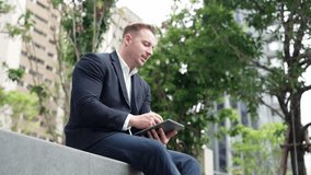Side view of businessman sitting at park while writing on tablet by using tablet pencil. Low angle camera of smart project manager looking at data analysis while thinking about financial plan. Urbane - Powered by Shutterstock - Get 15% off with code: PIKWIZARD15