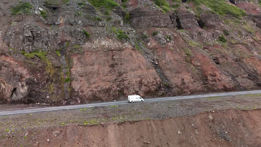 A white van navigates a scenic roadway along dramatic cliffs in Iceland during twilight. The landscape features rugged terrain, vibrant colors, and natural beauty, perfect for adventure seekers.
