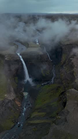 Witness Haifoss waterfall flowing through a deep canyon in Iceland, surrounded by lush greenery and dramatic rock formations. Mist envelops the scene, enhancing the beauty of nature.