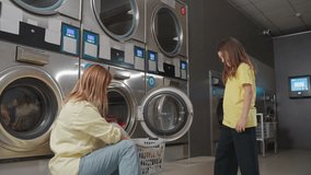 Mother with kid packing clothes into washer at laundromat, woman kneels by open stainless machine, child gestures while basket of towels waits, sorting textiles, clean service routine inside - Powered by Shutterstock - Get 15% off with code: PIKWIZARD15