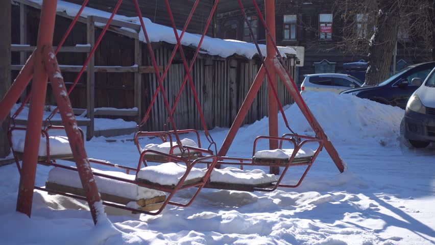 Melancholic swinging steel playground swing in empty winter courtyard. Typical poor Soviet-era yard with old authentic wooden log houses in background. Evokes loneliness, sadness and past memories 4k