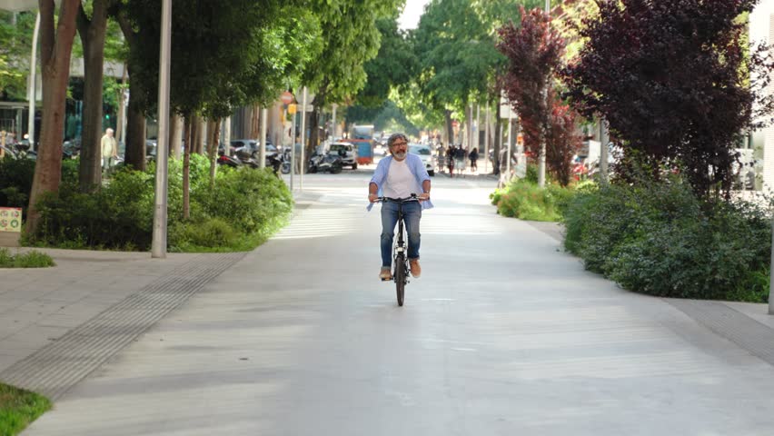 Senior man with glasses and a beard riding a folding bike in green city park street. Sustainable urban transportation