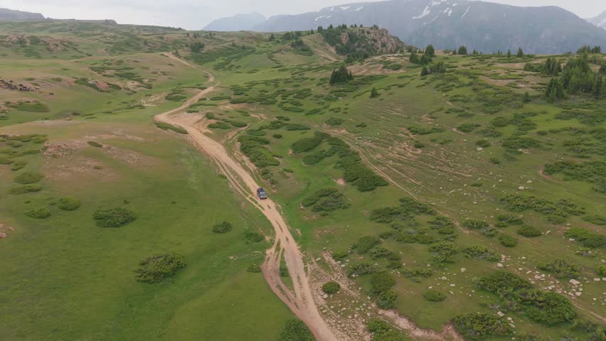 Rugged suv traversing winding dirt road through verdant meadows, rolling hills leading toward majestic snow capped mountain peaks in remote wilderness landscape