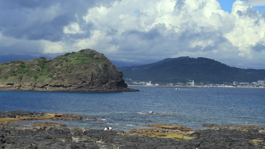 A wide view of Yongmeori Coast shows its tiered rock shelves and visitors along the waterline, with the town and low green hills across the bay under towering clouds.