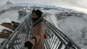 A woman is taking a selfie on a metal railing at the top of a mountain. The railing is surrounded by snow and the woman is wearing a scarf - Powered by Shutterstock - Get 15% off with code: PIKWIZARD15