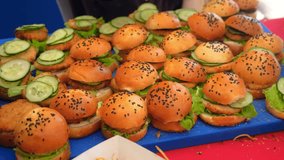 Chef cooking mini burgers adding halves of buns with black sesame seeds. Cook wearing black gloves preparing burgers with meat, salad and vegetables. Catering, street fast food. Unhealthy snack. - Powered by Shutterstock - Get 15% off with code: PIKWIZARD15